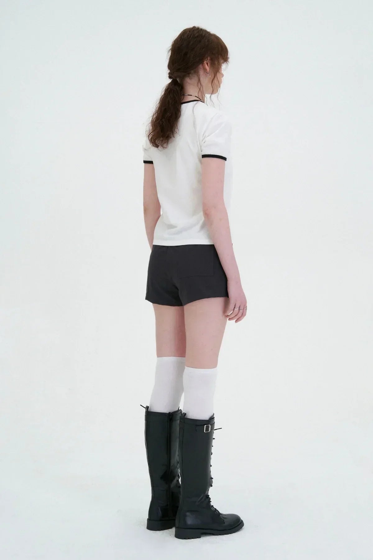 Woman wearing black shorts, white ringer t-shirt, knee-high white socks, and black lace-up boots on white background