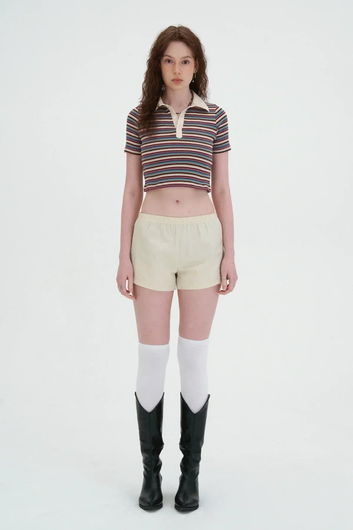 Woman in striped crop top, beige shorts, white knee socks, and black boots on white background