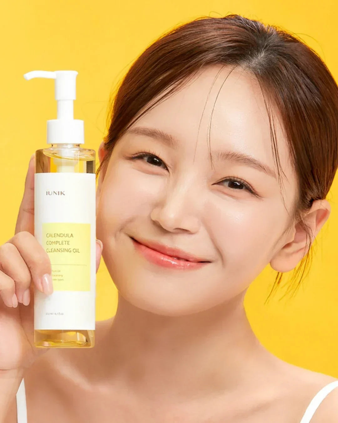 Smiling woman holding iUNIK Calendula Complete Cleansing Oil bottle with yellow background