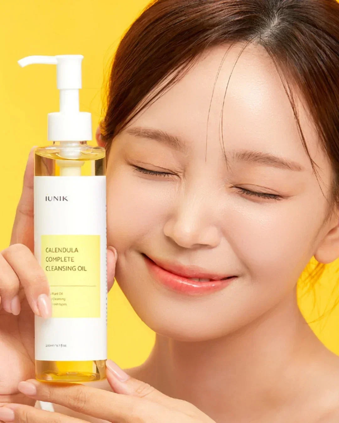 Smiling woman holding IUNIK Calendula Complete Cleansing Oil bottle with yellow background