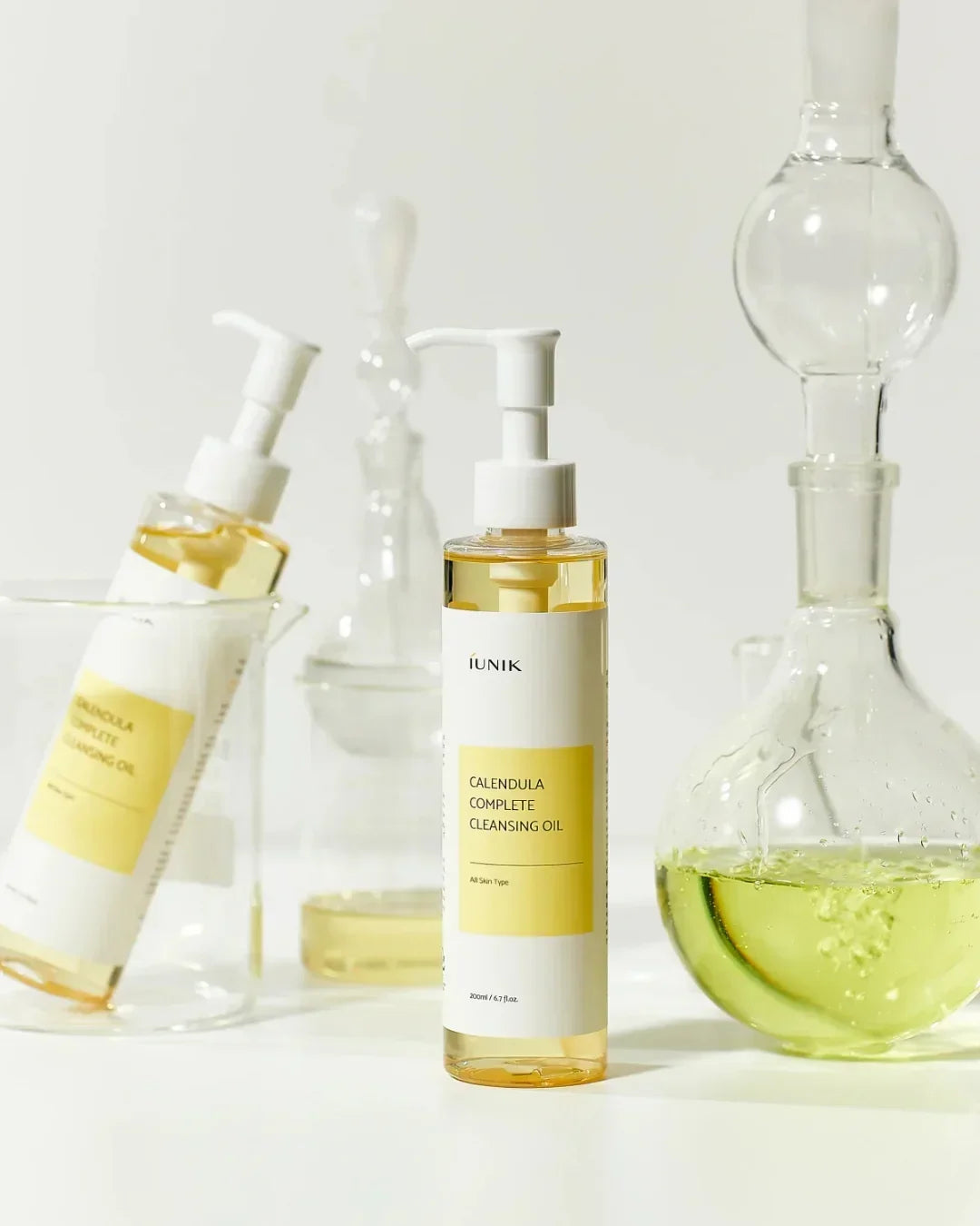 iUNIK Calendula Complete Cleansing Oil bottle with clear scientific glassware filled with yellow liquid on white background