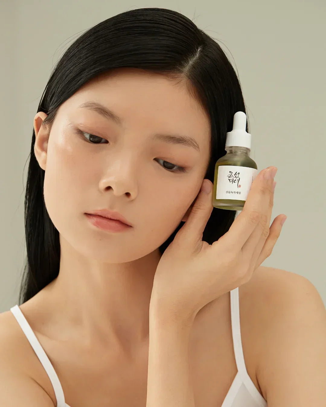 Young woman with clear skin holding green tea calming serum skincare bottle near her face