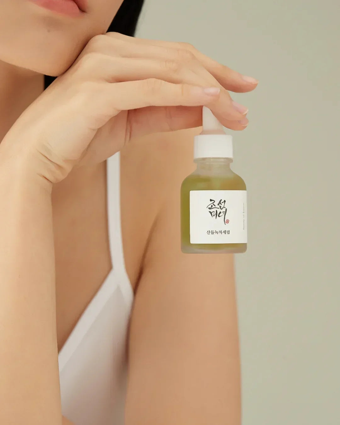 Woman holding a green calming serum bottle with white label and dropper against a neutral background