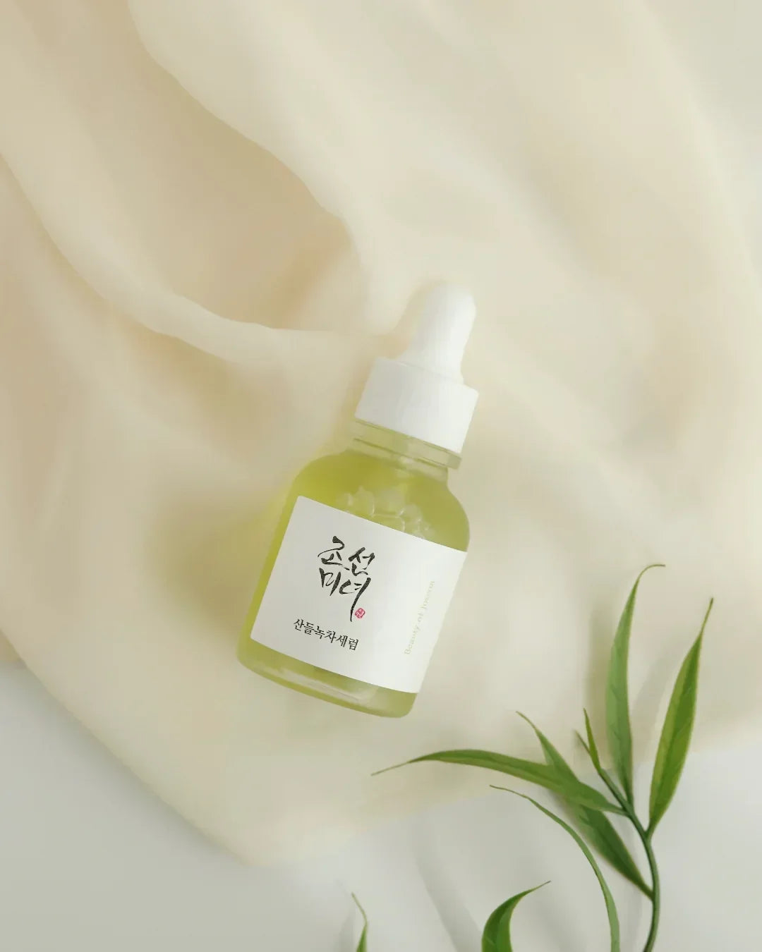 Green tea calming serum bottle with white dropper on beige fabric background and green leaves