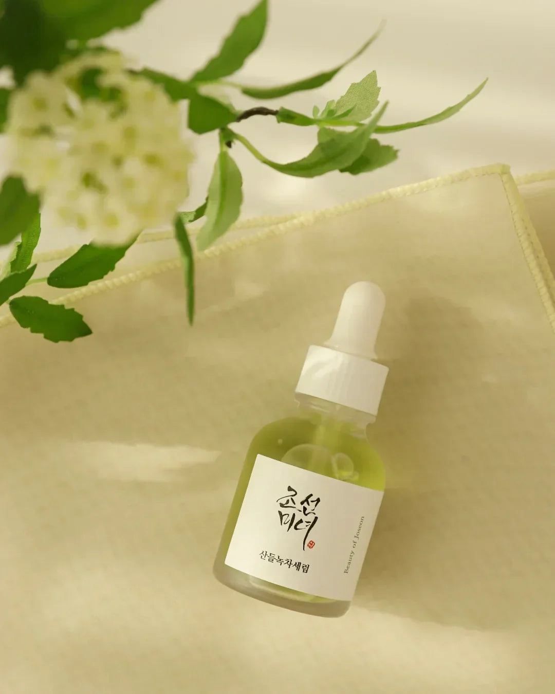 Green tea calming serum bottle with dropper on beige textured fabric and blurred white flower