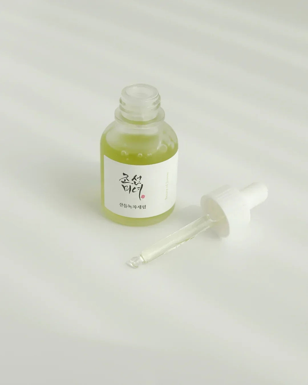 Glass bottle of green tea calming serum with dropper on white surface and soft shadows