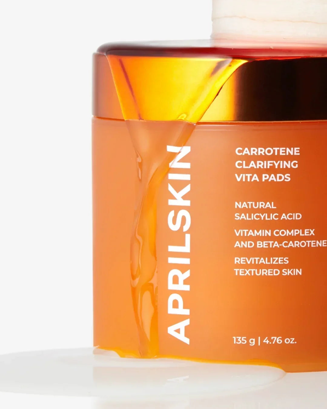 Aprilskin Carotene Clarifying Vita Pads jar with orange gel spilling over golden lid on white surface