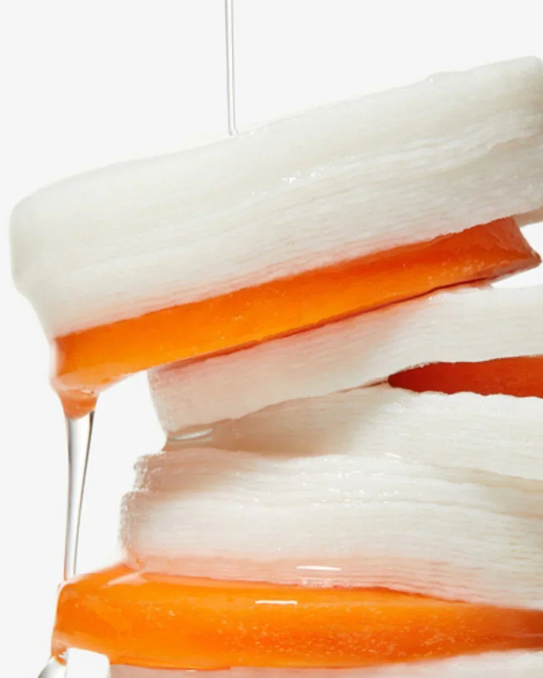 Close-up of stacked white cotton pads soaked with orange liquid serum dripping over edges