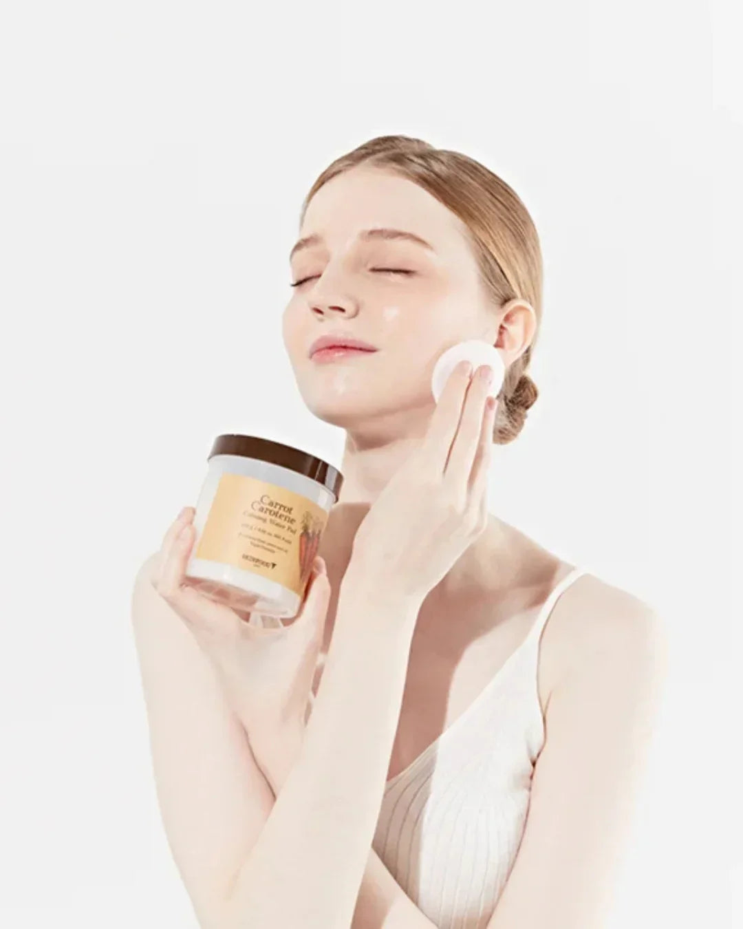 Young woman applying carrot carotene calming water pad on face holding the jar in a white top