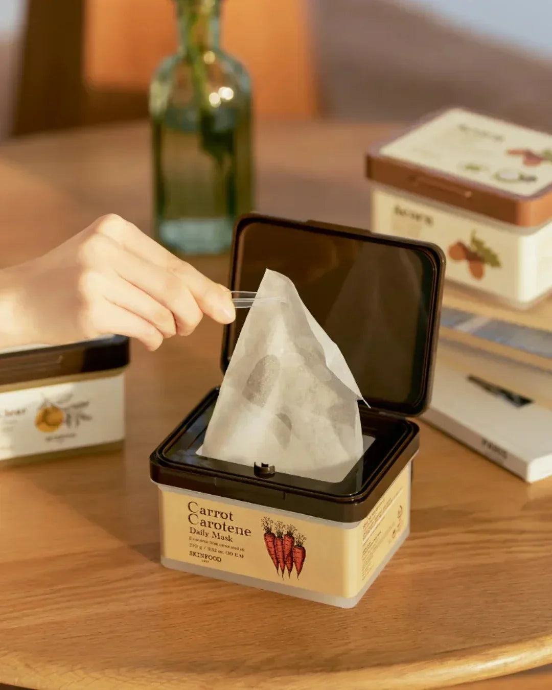 Hand pulling a sheet mask from a Skinfood Carrot Carotene Daily Mask container on a wooden table