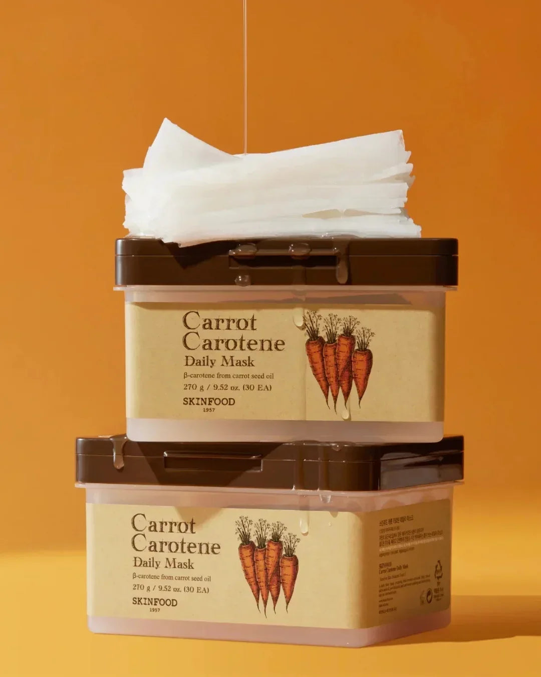 Stacked Skinfood Carrot Carotene Daily Mask containers with wet brown lids and white mask sheets on orange background