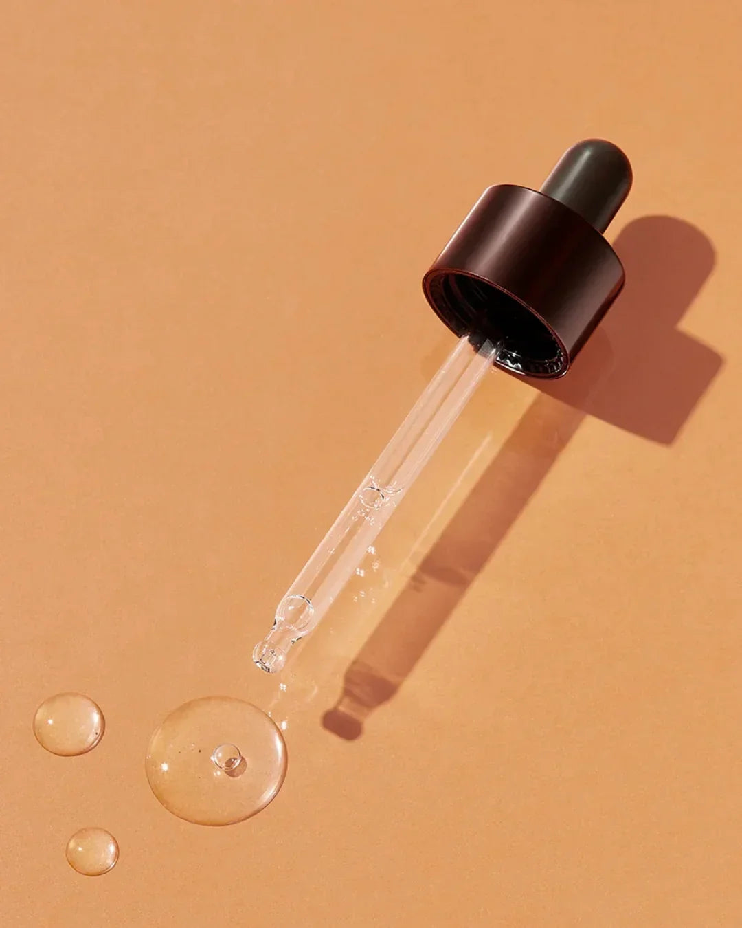 Close-up of a glass dropper with liquid droplets on a peach-colored surface, skincare serum texture