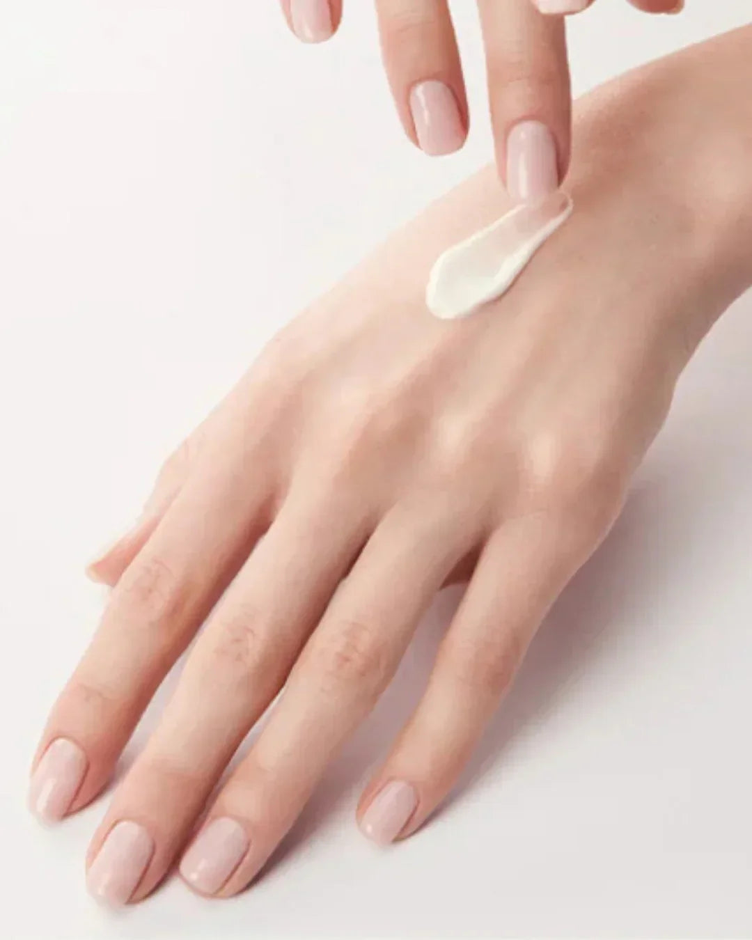 Close-up of hand with light pink polished nails applying white moisturizing cream on the back of the hand