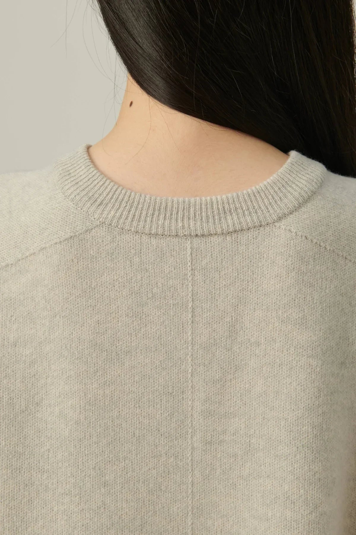 Close-up of beige cashmere crewneck sweater on person with dark hair against neutral background