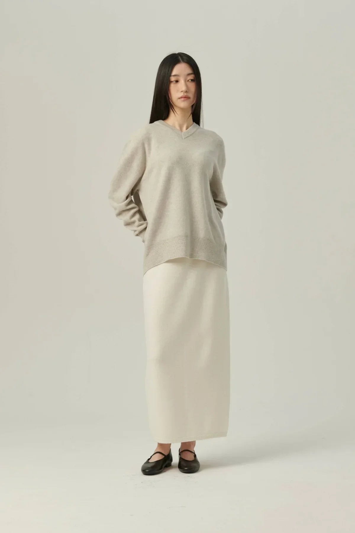 Woman wearing beige cashmere V-neck pullover and long cream skirt with black shoes on white background