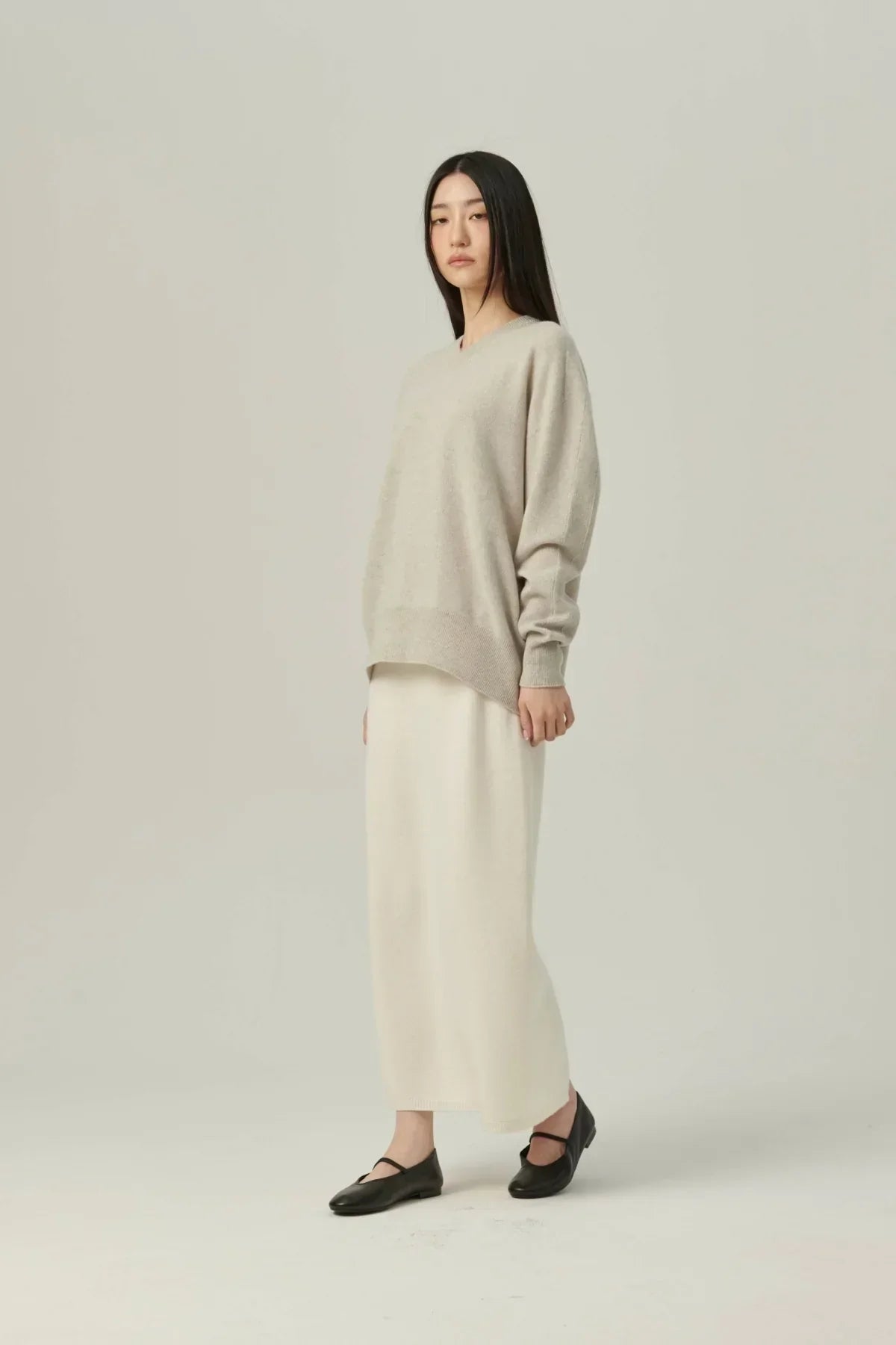 Woman wearing beige cashmere V-neck pullover with long cream skirt and black flat shoes on plain background
