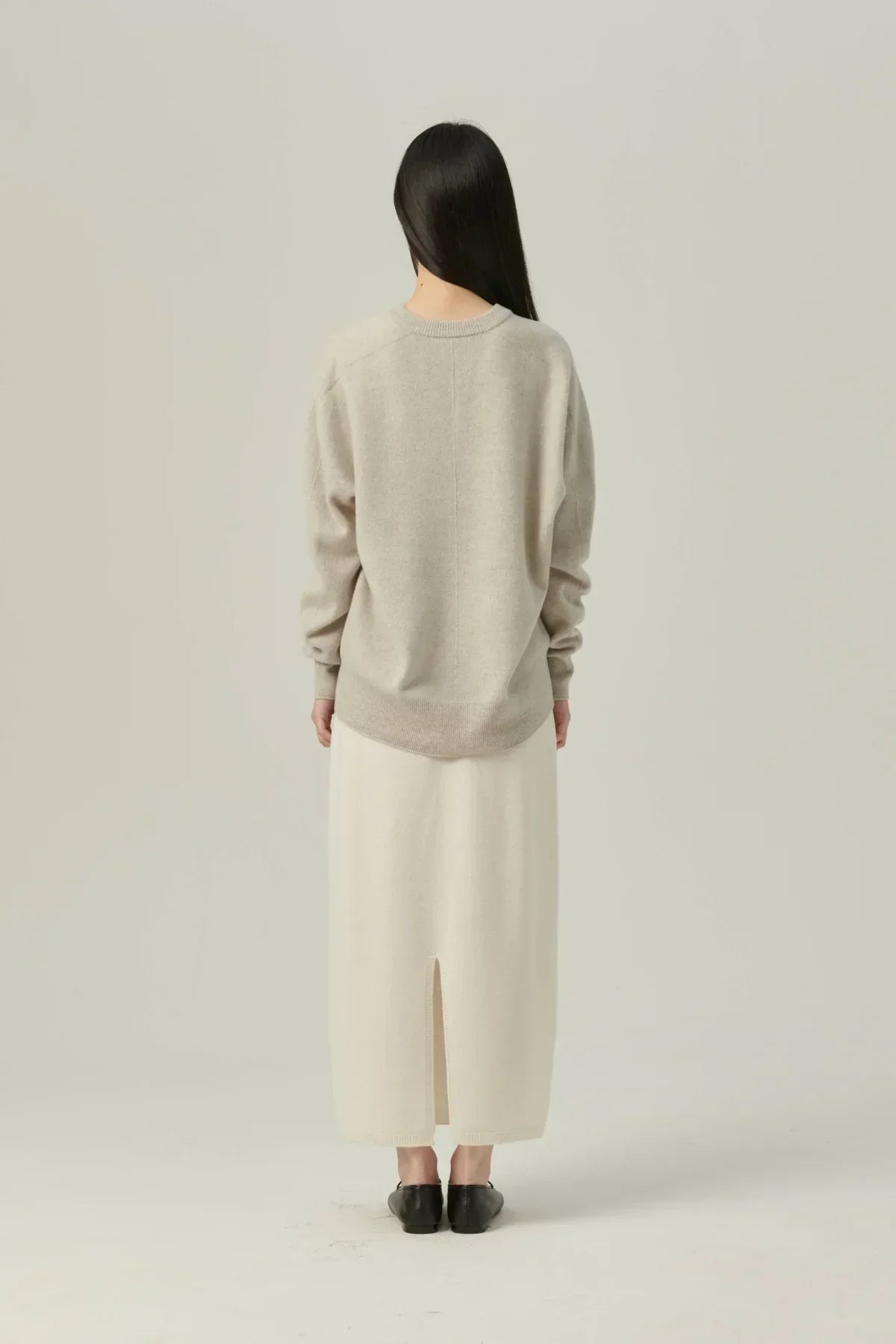 Woman wearing beige cashmere pullover and cream long skirt with back slit, black shoes, minimal studio background