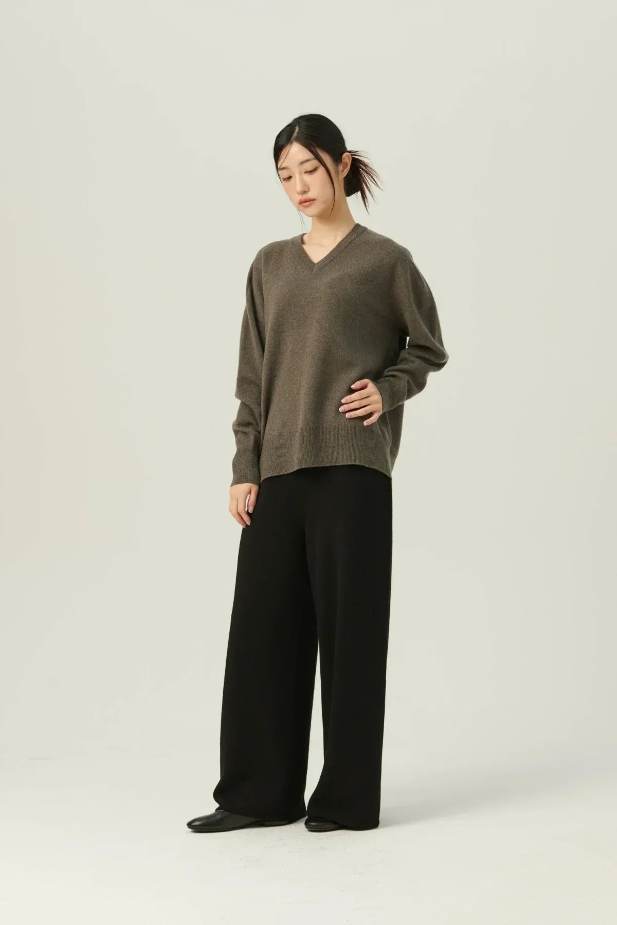woman wearing brown cashmere V-neck pullover and black wide-leg pants standing against light background