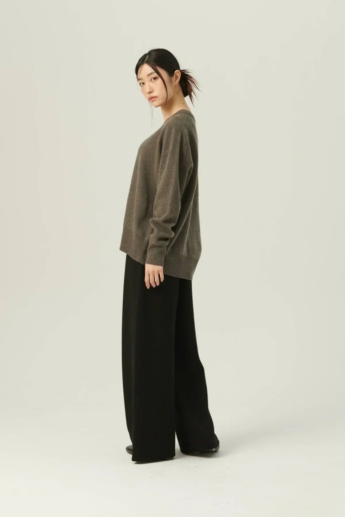 Side view of woman wearing brown cashmere V-neck sweater and black wide-leg pants on plain background