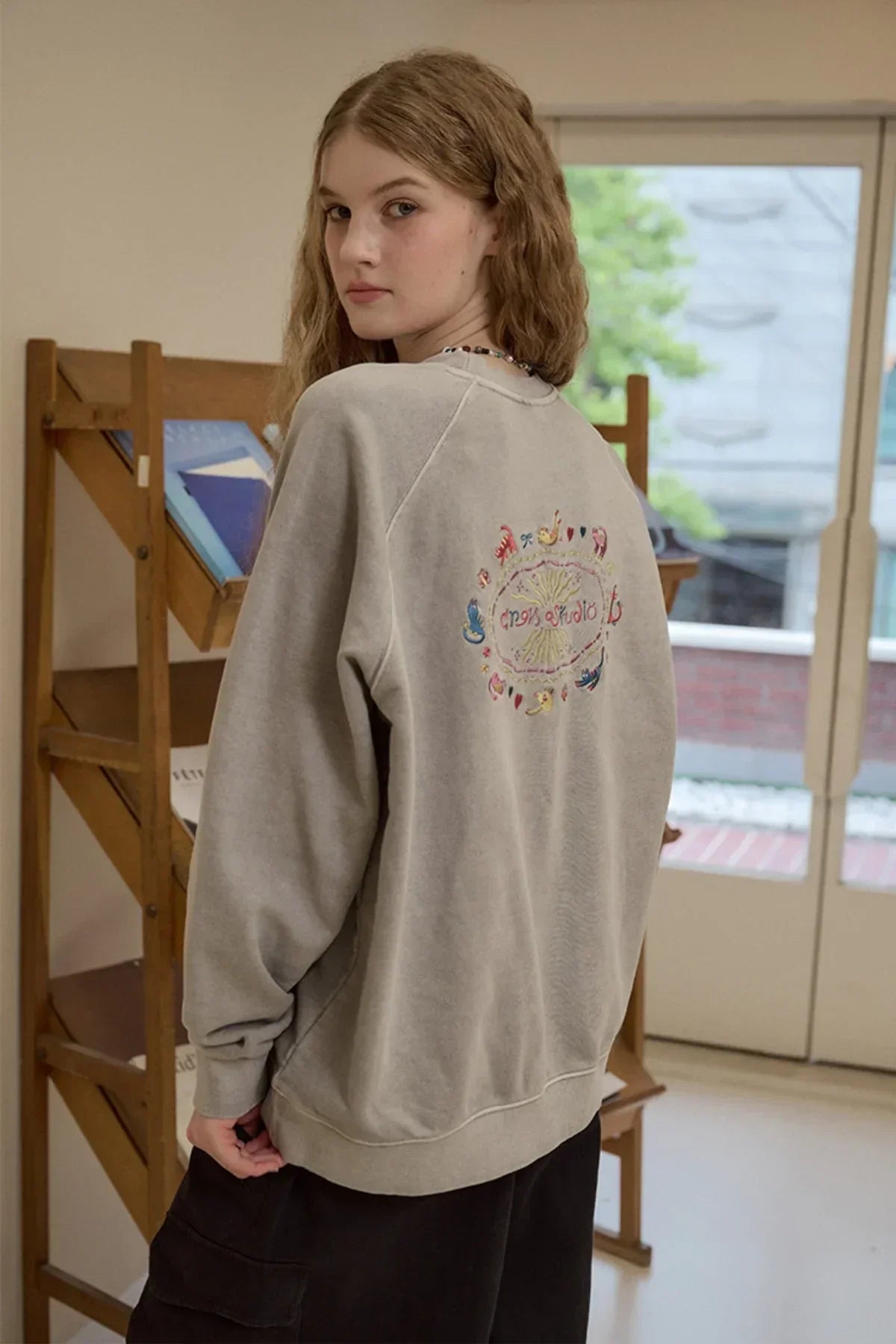 Young woman indoors wearing ghost gray raglan sweatshirt with colorful cat embroidery on back