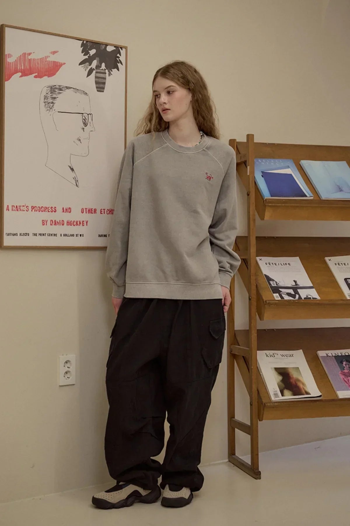 Woman wearing gray sweatshirt and black cargo pants standing near wooden shelf with magazines and framed artwork