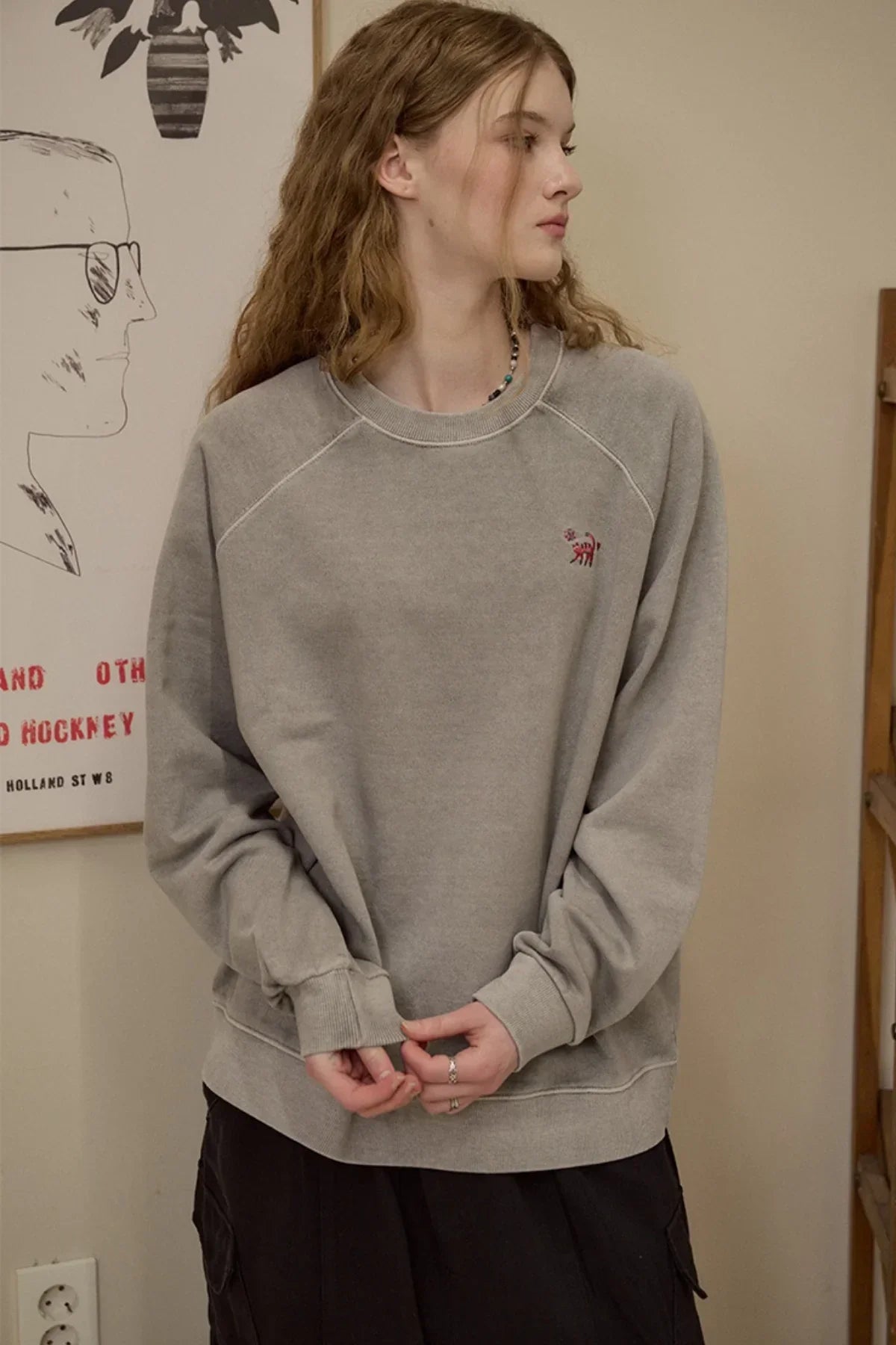 Woman wearing gray raglan sweatshirt with small cat embroidery on chest in indoor setting