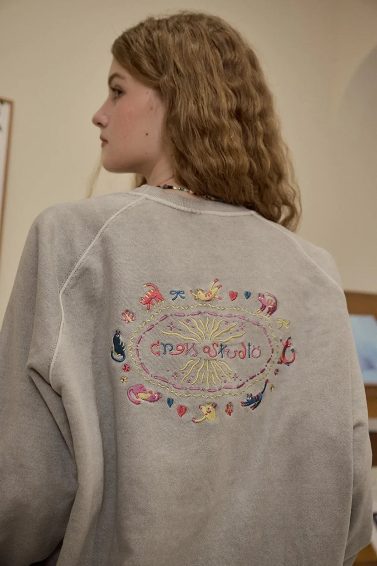 Woman wearing ghost gray raglan sweatshirt with colorful embroidered cat design on back
