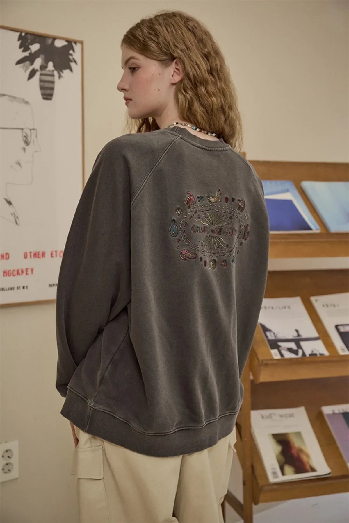 Woman wearing a charcoal grey oversized sweatshirt with embroidered back design in a cozy room with wooden magazine rack