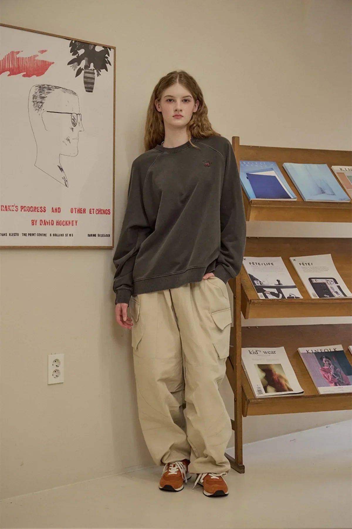 Young woman in black oversized sweatshirt and beige cargo pants standing indoors by a magazine rack and framed wall art