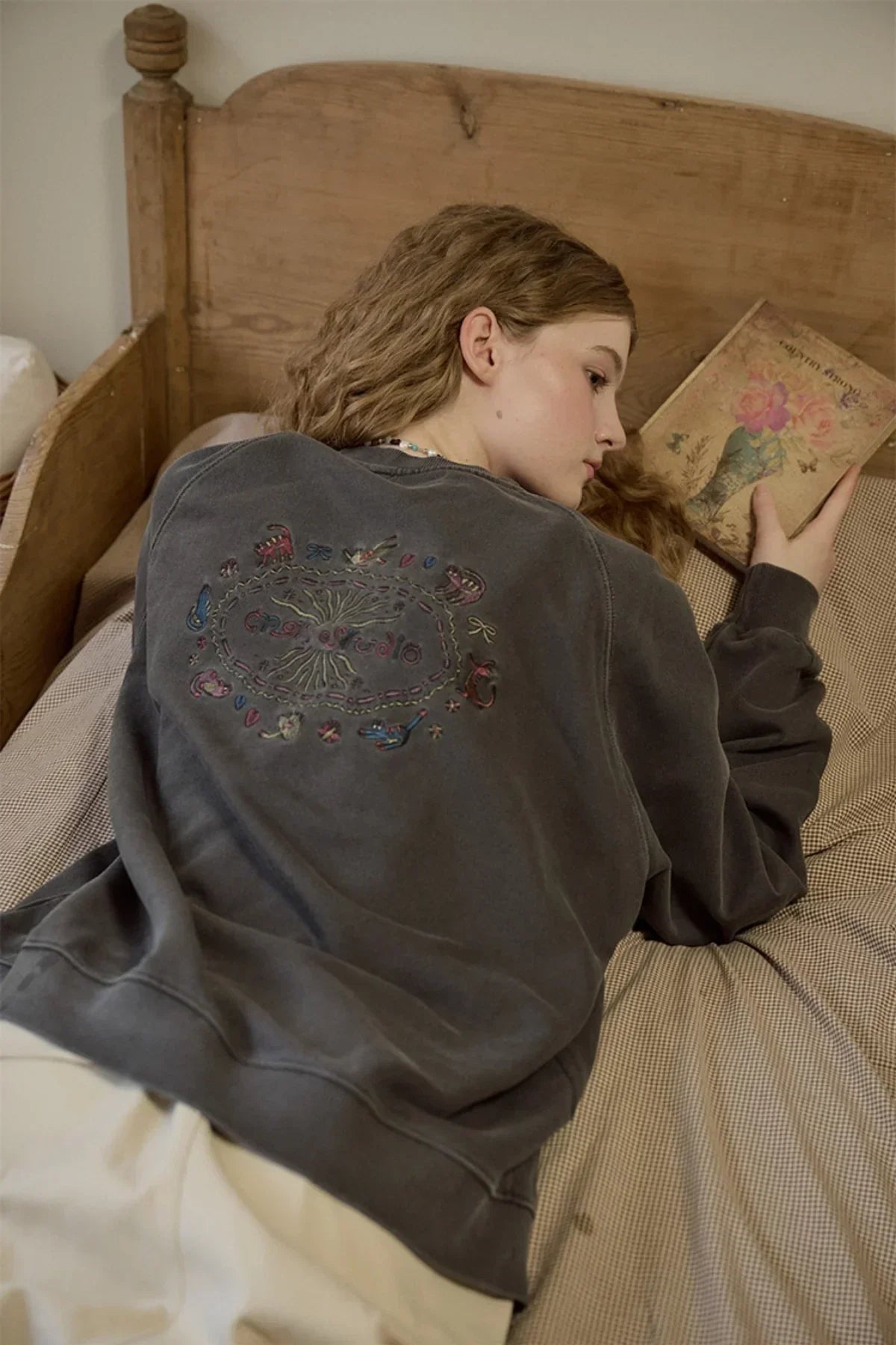Woman lying on wooden bed reading book, wearing dark gray sweatshirt with colorful embroidered back design
