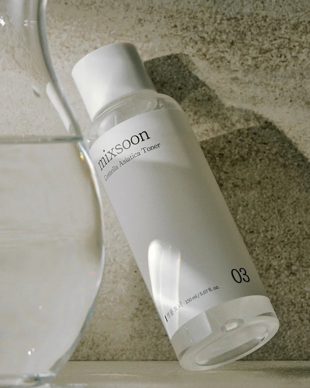 Mixsoon Centella Asiatica Toner 150ml bottle on textured beige surface with glass of water