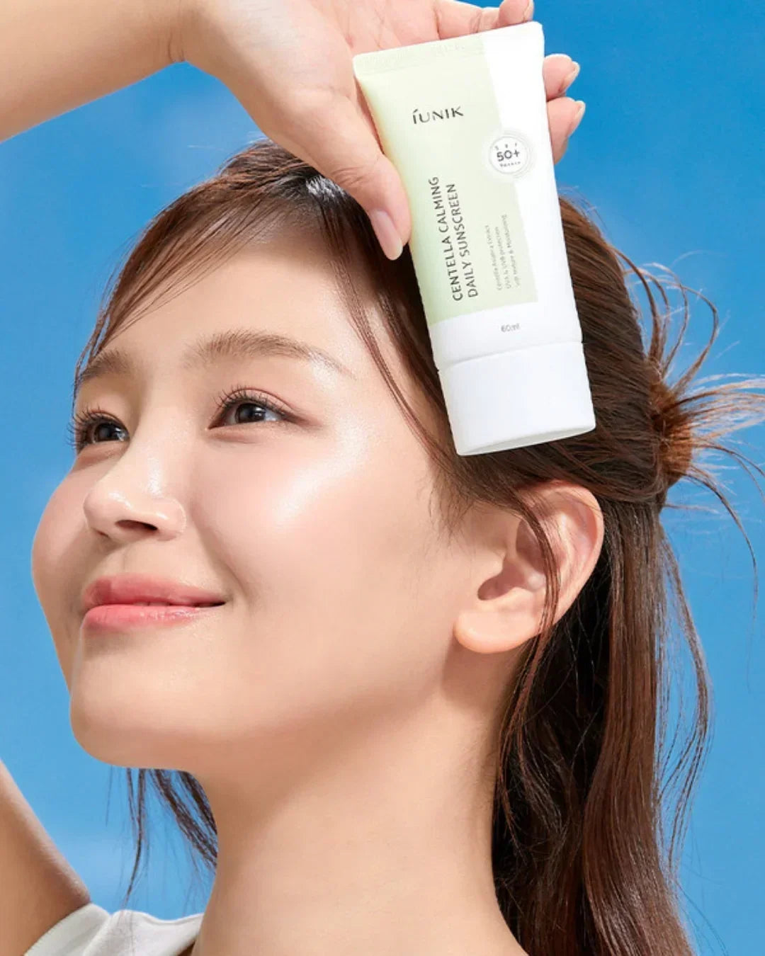 Young woman with clear skin holding iUNIK Centella Calming Daily Sunscreen SPF 50+ tube against blue background