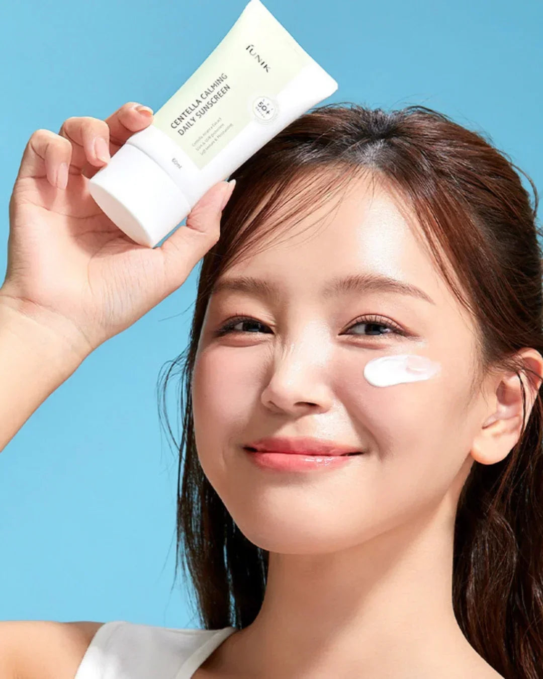 Smiling woman applying Centella Calming Daily Sunscreen with SPF 50+ on cheek against blue background