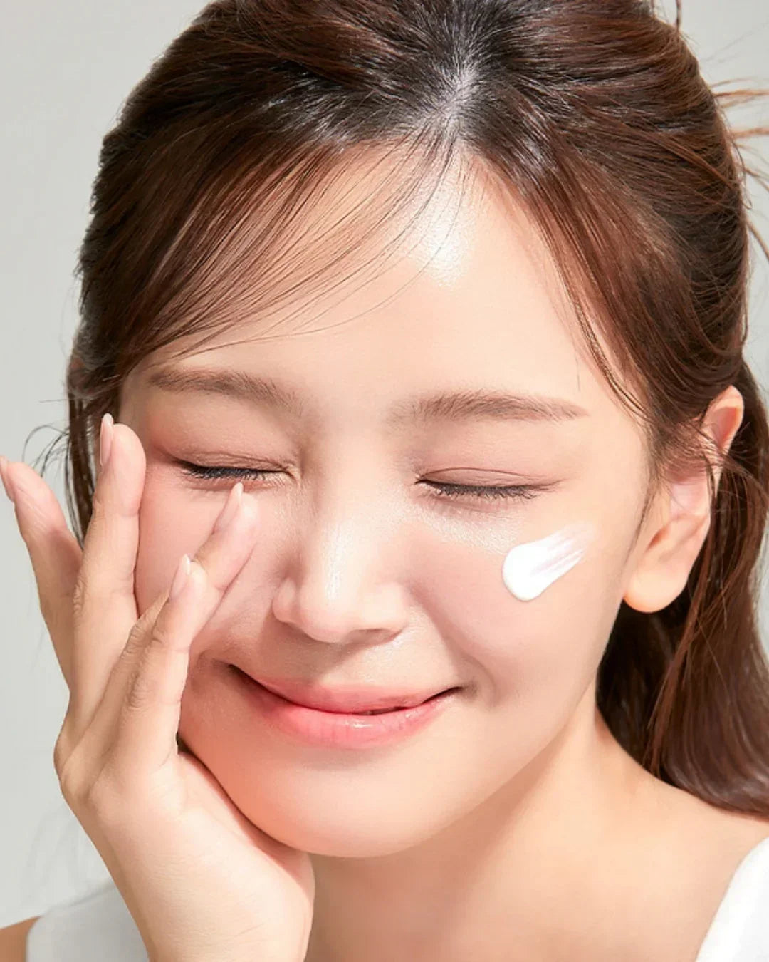 Close-up of woman applying white sunscreen cream on cheek with closed eyes and smiling