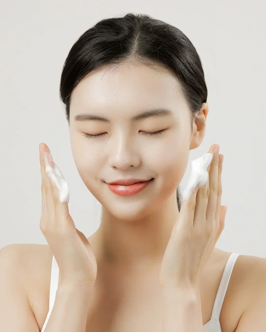 Young woman with clear skin applying white cleansing foam on cheeks with closed eyes