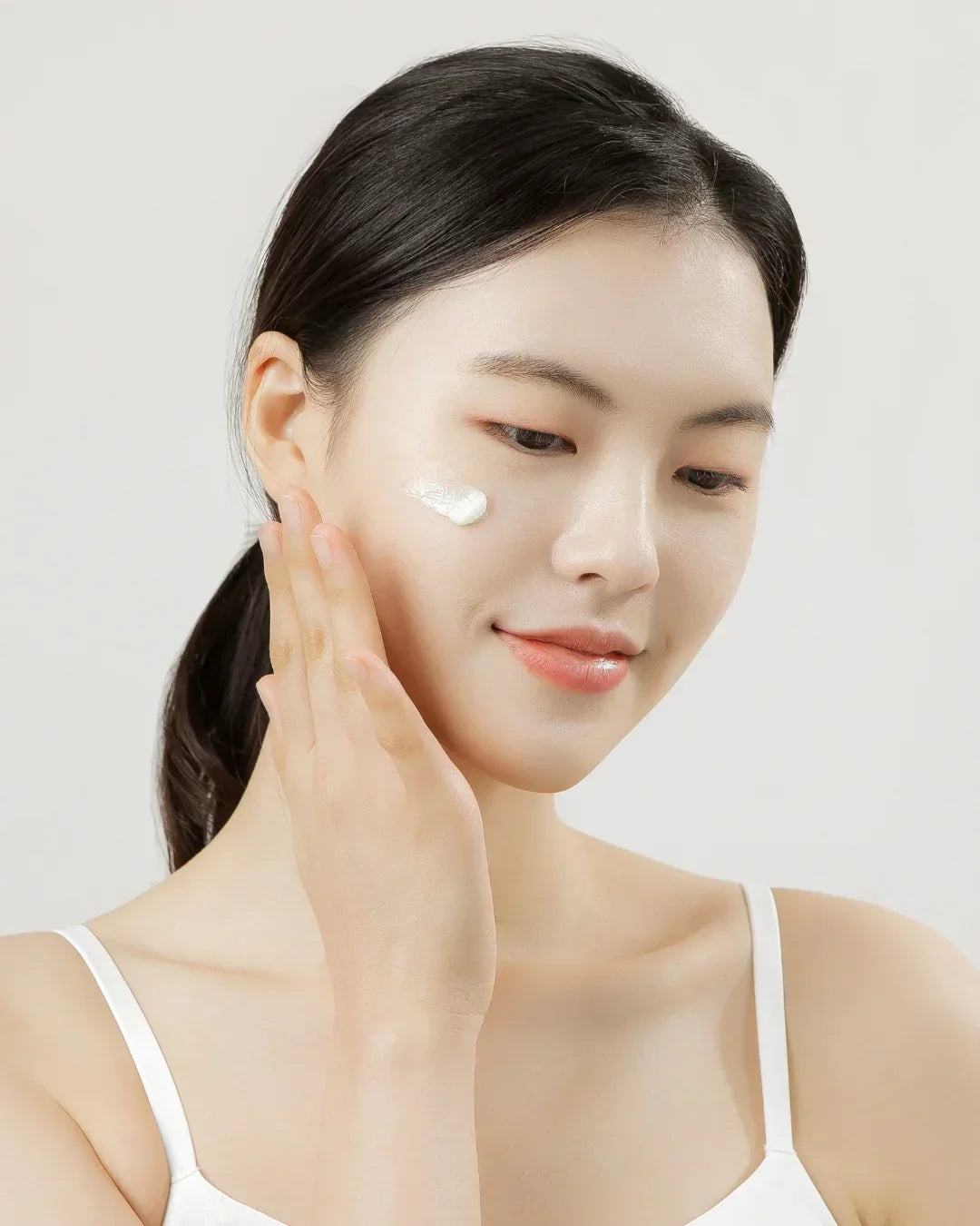 Young woman with clear skin applying white cream on cheek, skincare beauty routine