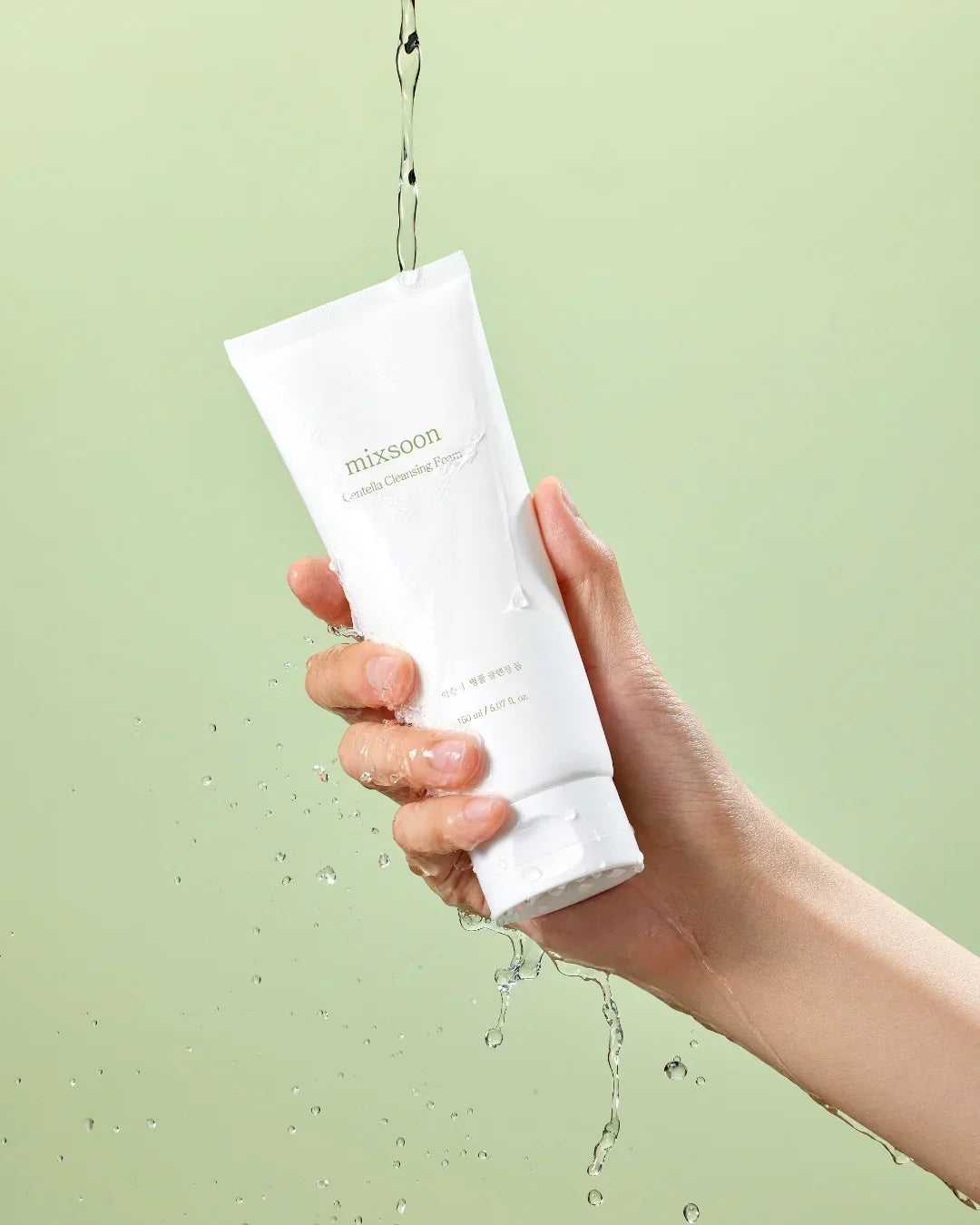 Hand holding Mixsoon Centella Cleansing Foam tube with water splashing on green background