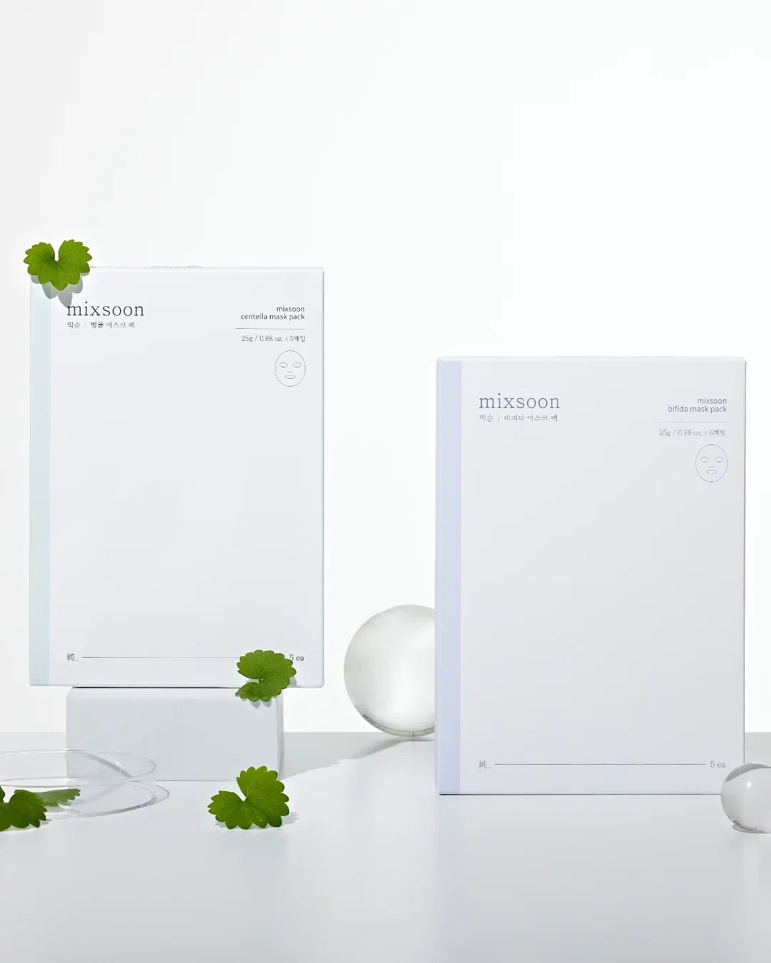 Two white mixsoon face mask pack boxes with green centella leaves and clear glass spheres on white surface
