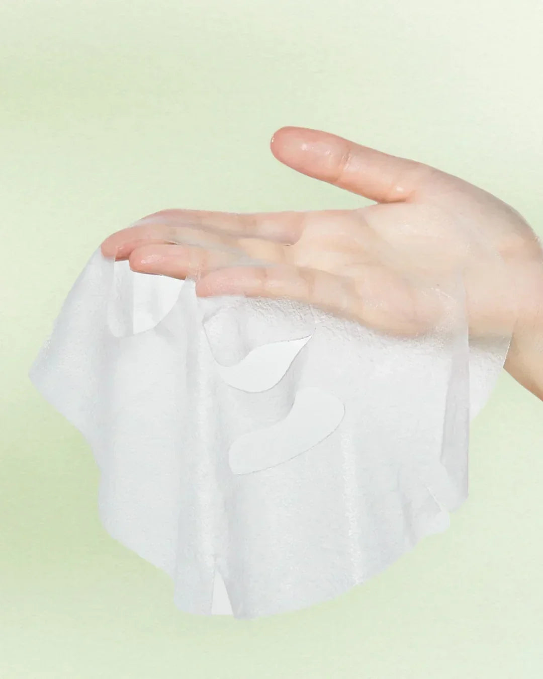 Hand holding wet translucent sheet face mask on light green background