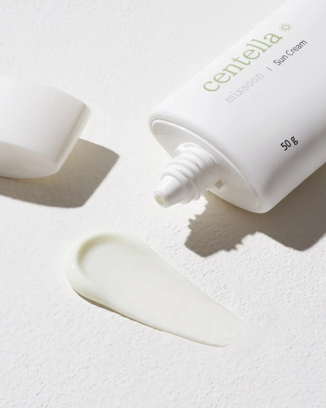 White tube of Centella sun cream with cap removed and cream swatch on textured white surface
