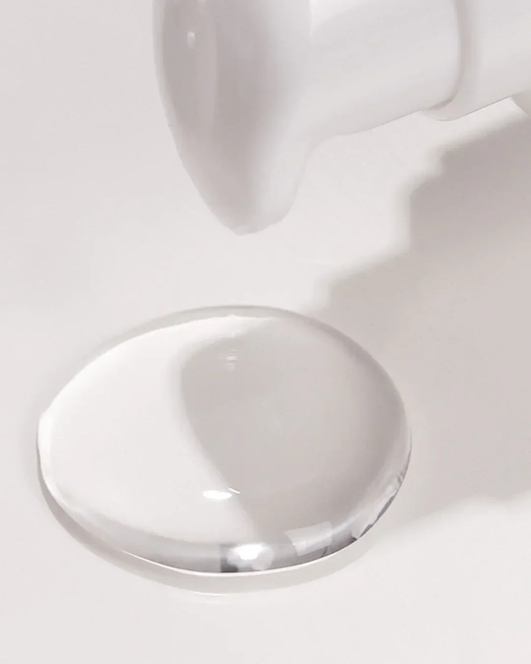 Clear, glossy essence drop on a white surface with a white bottle nozzle above