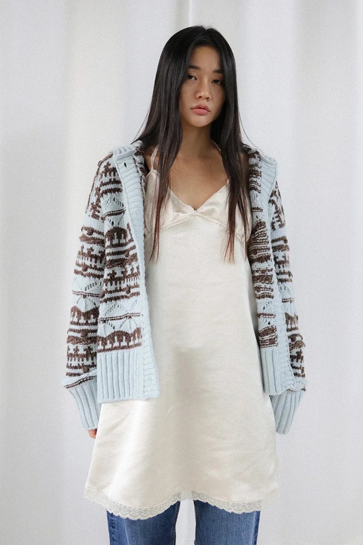 Woman wearing a light blue chunky knit cardigan with brown pattern over a cream satin slip dress and blue jeans