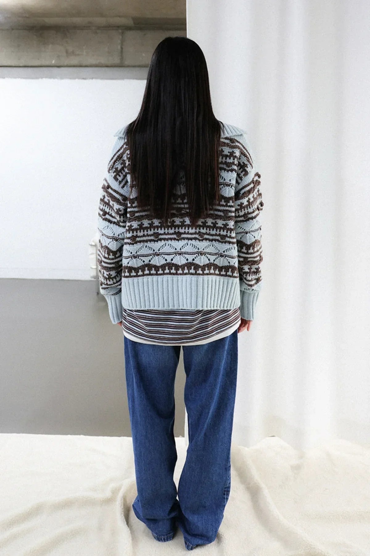 Woman wearing chunky knit patterned cardigan over striped shirt and loose blue jeans, back view