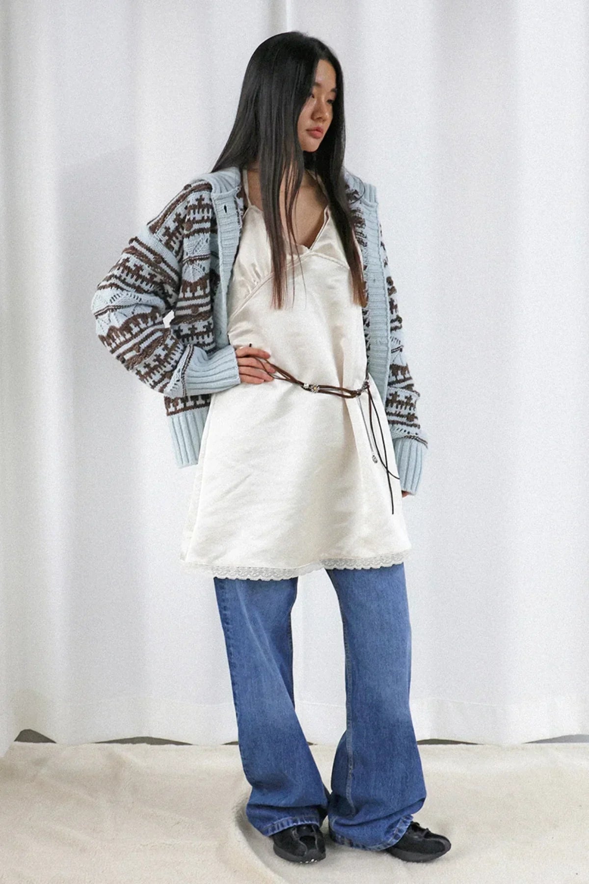 Young woman wearing chunky knit cardigan, satin slip dress with brown belt, blue jeans, and black shoes