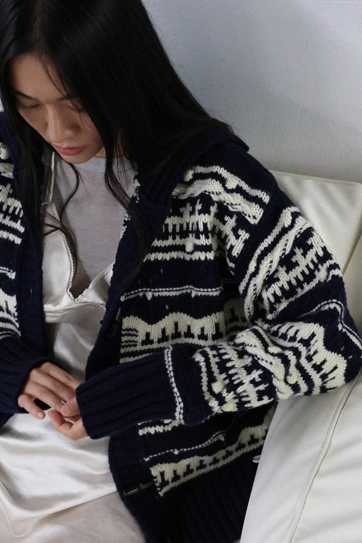 Woman wearing navy chunky knit cardigan with white patterns, sitting on white couch