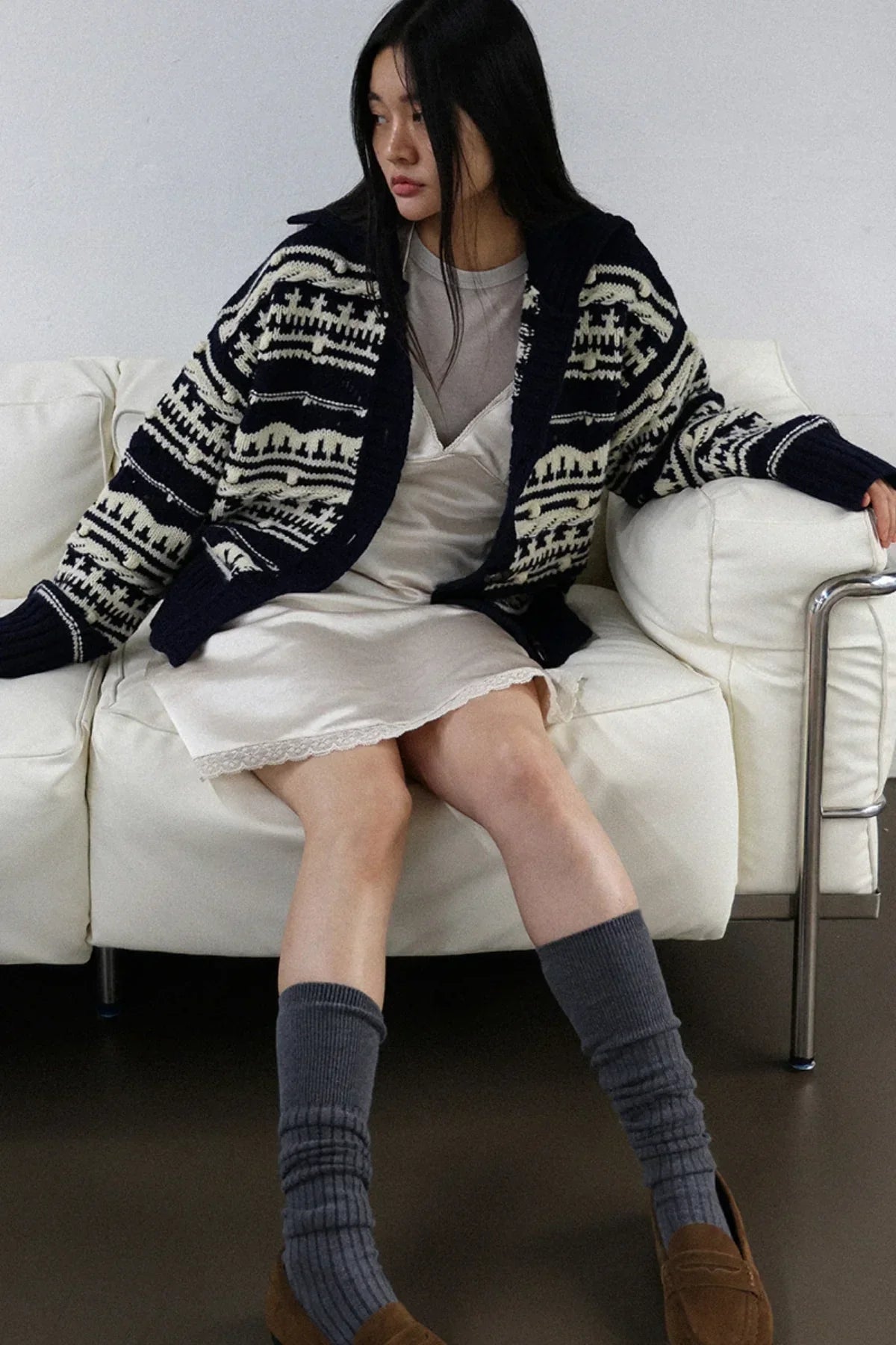 Woman sitting on white sofa wearing navy chunky collar box fit cardigan, cream slip dress, gray socks, and brown loafers