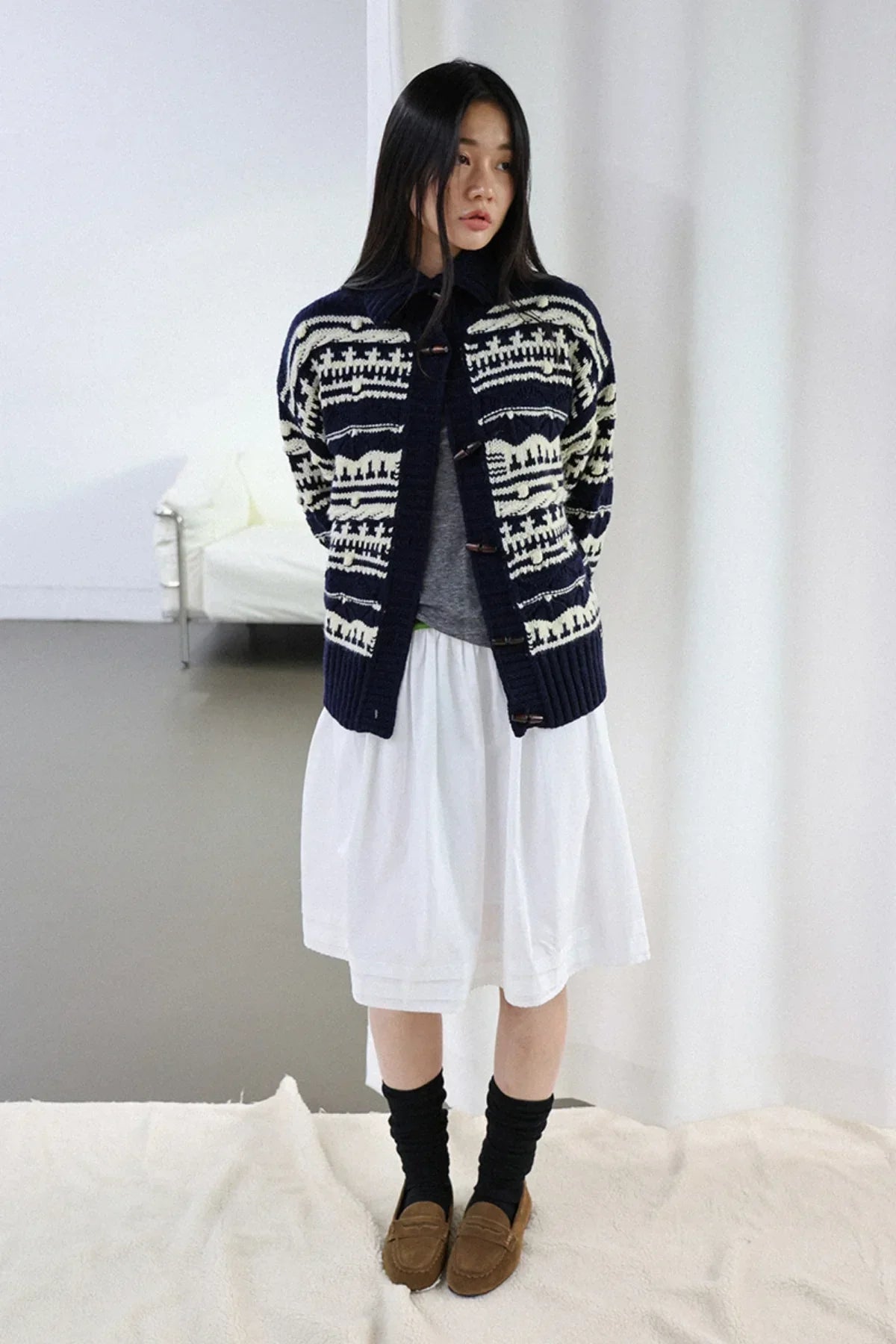 Woman wearing navy chunky collar box-fit cardigan with white pattern over gray top and white skirt indoors
