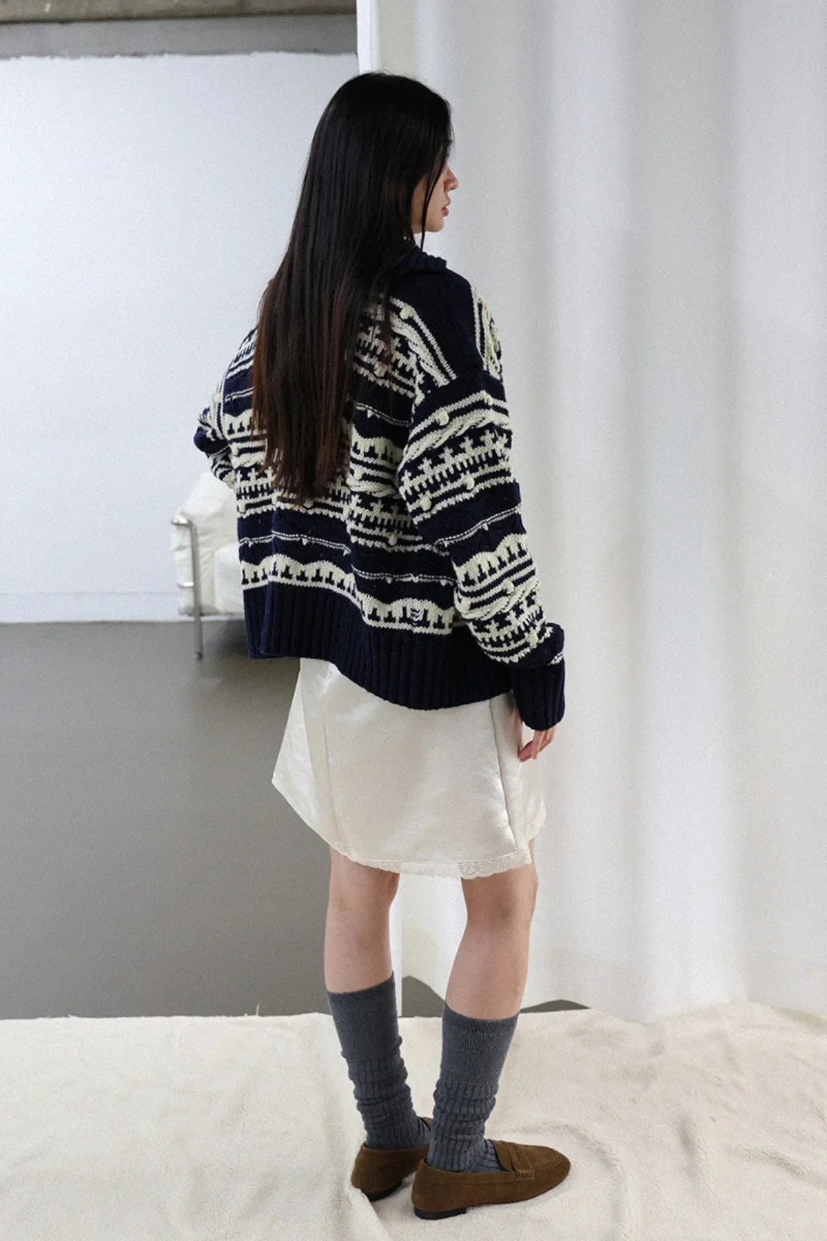 Woman wearing navy and white patterned chunky knit cardigan with white skirt, gray socks, and brown loafers