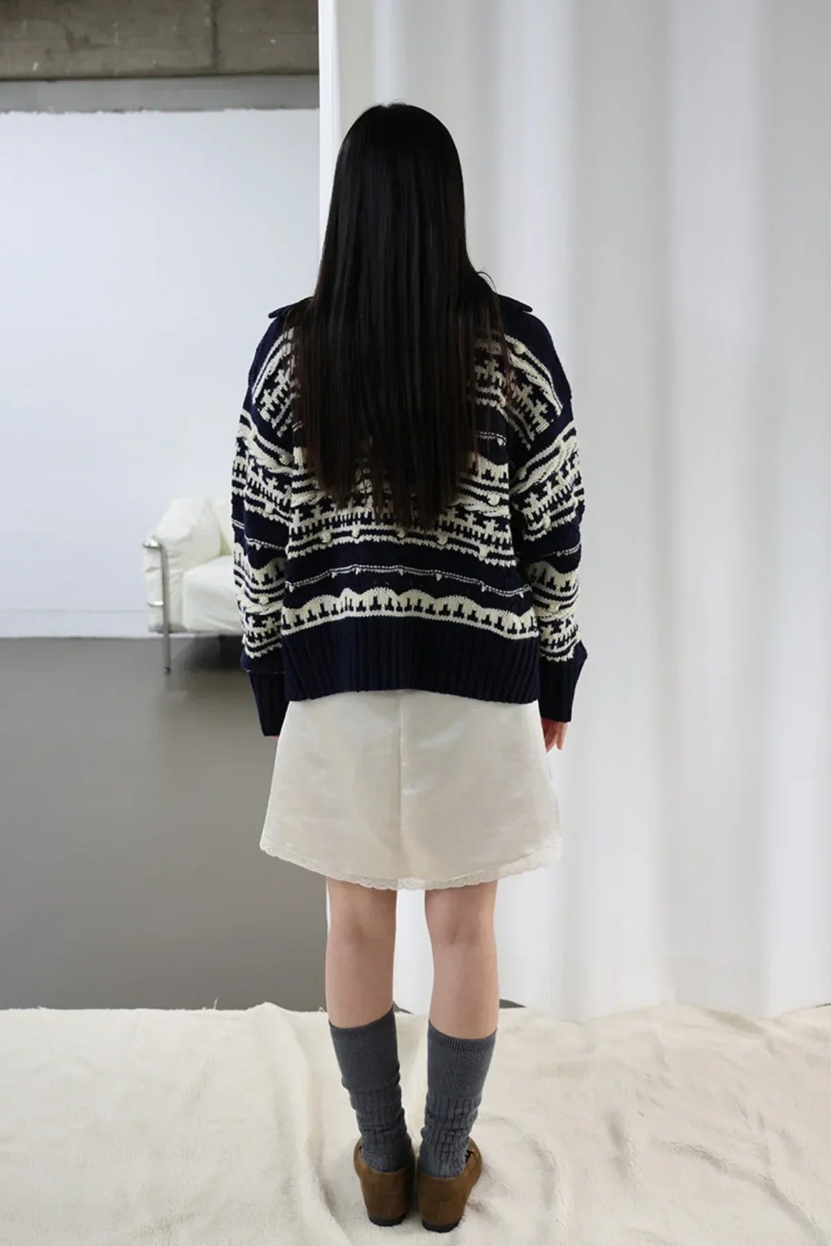 Back view of woman wearing navy chunky collar box fit cardigan with white pattern and cream skirt indoors