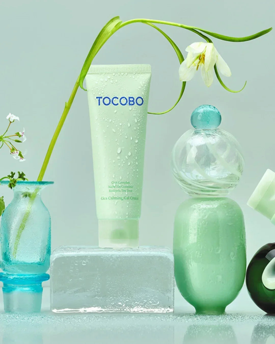 TOCOBO Gica Calming Gel Cream tube with water droplets, surrounded by green vases and white flowers